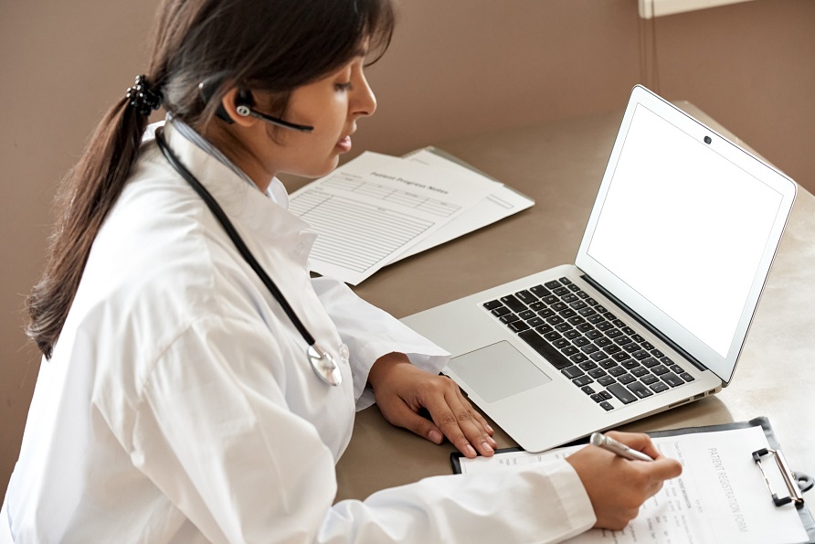 Woman in doctors coat writes on clipboard sitting in front of laptop